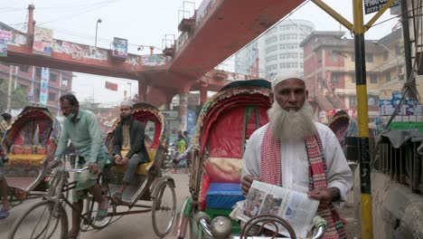 Un-Hombre,-Un-Tirador-De-Rickshaw,-Lee-Un-Periódico-Al-Aire-Libre-Mientras-Los-Viajeros-Conducen-Y-Pasan-A-Través-Del-Marco-En-Una-Calle-Concurrida