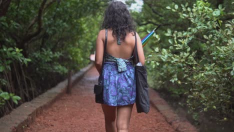 Dark-skin-woman-walks-in-blue-dress-through-park-path,-holds-umbrella-slow-motion