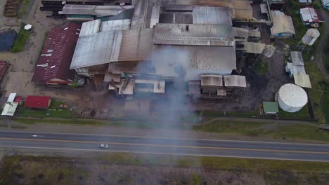 Aerial-drone-shot-of-a-factory-near-a-palm-plantation,-overhead-shot-of-the-building-roof-with-a-steaming-chimney