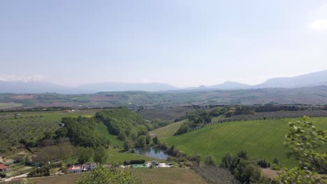 Drone-shot-going-through-trees-and-branches-revealing-a-wide-open-scene-of-a-valley-with-farmland,-wineries-and-mountains-in-the-distance-in-the-countryside-of-Abruzzo-in-Italy