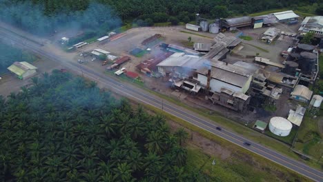 A-factory-near-a-palm-plantation,-aerial-drone-shot-of-the-building-roof-with-a-steaming-chimney,-rotating-tracking-movement