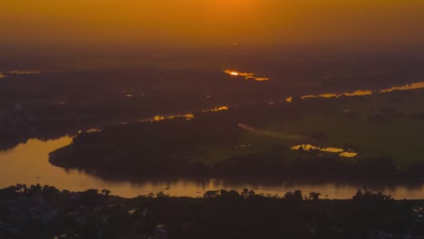 Timelapse-of-a-golden-sunset-over-a-foggy-rural-countryside,-with-small-boats-sailing-down-the-river