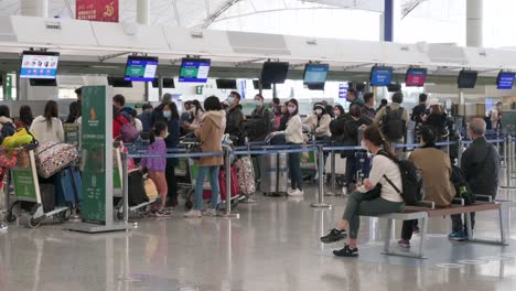 Chinesische-Flugpassagiere-Sitzen-Auf-Einer-Bank,-Während-Andere-Reisende-Am-Schalter-Der-Fluggesellschaft-Im-Hintergrund-Am-Internationalen-Flughafen-Hongkong-Einchecken
