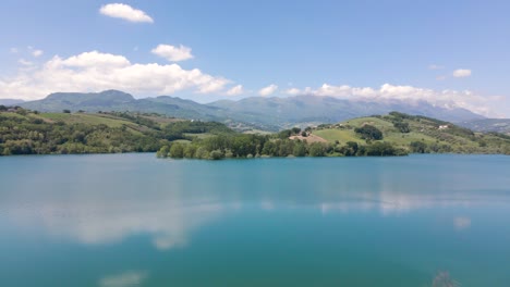 Wide-angle-drone-shot-revealing-a-tranquil-scene-of-a-beautiful-emerald-lake-surrounded-by-mountains-in-a-valley-near-the-small-village-of-Penne,-in-the-region-of-Abruzzo,-Italy