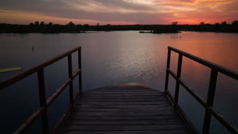 Beautiful-orange-and-pink-sunset-tilt-up-from-old-wooden-dock-looking-out-over-lake-with-colors-reflecting-off-water
