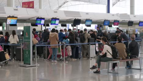 Flugpassagiere-Sitzen-Auf-Einer-Bank,-Während-Andere-Reisende-Am-Schalter-Der-Fluggesellschaft-Im-Hintergrund-Am-Internationalen-Flughafen-Hongkong-Einchecken