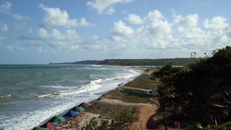 Tilt-up-of-Gramame-beach-during-high-tide-where-the-ocean-meets-the-river-with-waves-crashing-into-the-sand-and-colorful-umbrellas-blowing-in-the-wind-near-the-city-of-Joao-Pessoa-in-Paraiba,-Brazil