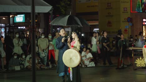 Dúo-Vietnamita-Cantando-Una-Canción-Bajo-Un-Paraguas-Por-La-Noche-Con-El-Público-Mirando-En-La-Calle-Nguyen-Hue-En-Ho-Chi-Minh,-Vietnam