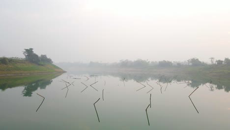 Aerial-footage-captures-a-foggy-morning,-flying-low-over-a-calm,-reflecting-big-river,-with-sticks-sticking-out-of-the-water