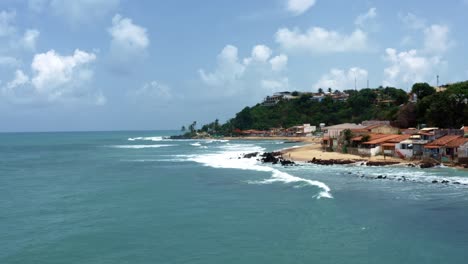 Dolly-in-truck-left-aerial-drone-wide-shot-of-the-tropical-famous-Baia-Formosa-beach-town-in-the-state-of-Rio-Grande-do-Norte,-Brazil-with-fishing-boats,-coastal-homes,-small-waves,-and-surfers