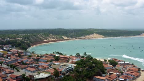 Truck-left-aerial-drone-wide-shot-flying-over-homes-to-the-famous-Baia-Formosa-beach-in-the-state-of-Rio-Grande-do-Norte,-Brazil-with-fishing-boats,-coastal-homes,-and-small-waves-on-a-summer-day