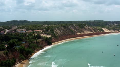 Dolly-in-aerial-drone-wide-shot-of-the-Cacimba-beach-cliffs-in-the-famous-beach-town-of-Baia-Formosa-in-Rio-Grande-do-Norte,-Brazil-with-fishing-boats-docked-and-small-waves-in-turquoise-water