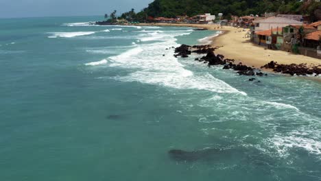 Dolly-in-tilt-up-aerial-drone-shot-of-the-tropical-famous-Baia-Formosa-beach-town-in-the-state-of-Rio-Grande-do-Norte,-Brazil-with-fishing-boats,-coastal-homes,-small-waves,-and-surfers