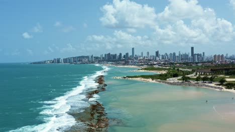 Dolly-out-aerial-shot-revealing-the-historic-star-shaped-Reis-Magos-fort-built-on-a-reef-with-the-coastal-capital-city-of-Natal-in-Rio-Grande-do-Norte,-Brazil-in-the-background-on-a-warm-summer-day