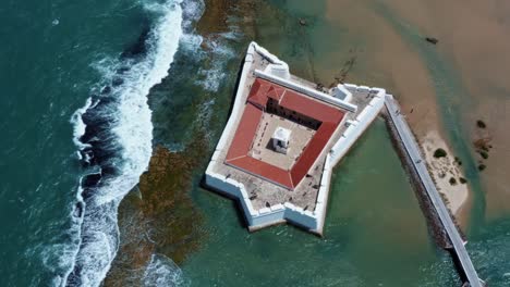 Tilting-up-aerial-drone-shot-of-the-historic-star-shaped-Reis-Magos-fort-built-on-a-reef-revealing-the-coastal-capital-city-of-Natal-in-Rio-Grande-do-Norte,-Brazil-in-the-background