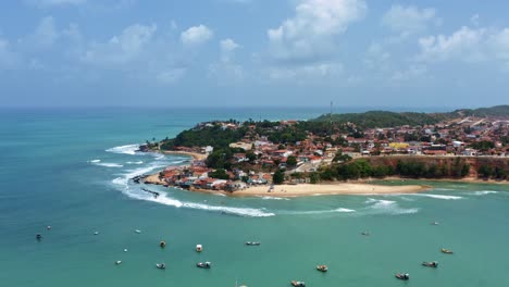 Tilt-down-aerial-drone-wide-shot-of-the-Cacimba-beach-in-the-beach-town-of-Baia-Formosa-in-Rio-Grande-do-Norte,-Brazil-with-fishing-boats,-coastal-houses,-small-waves,-and-sea-birds-flying-around