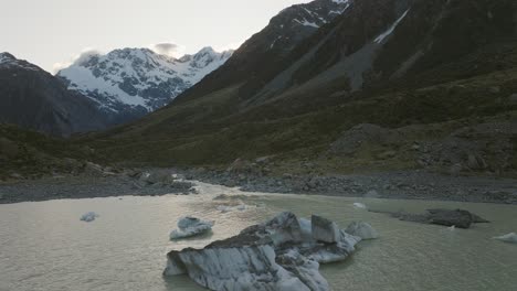Hooker-valley-glacial-lake-with-iceberg-and-tributary-flowing-downhill,-aerial