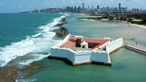 Dolly-in-aerial-drone-shot-of-the-historic-star-shaped-Reis-Magos-fort-built-on-a-reef-with-the-coastal-capital-city-of-Natal-in-Rio-Grande-do-Norte,-Brazil-in-the-background-on-a-warm-summer-day