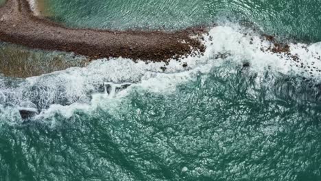 Descending-bird's-eye-aerial-drone-shot-of-tropical-turquoise-ocean-water-waves-crashing-into-a-man-made-breakwater-in-the-coastal-capital-city-of-Natal-in-Rio-Grande-do-Norte,-Brazil