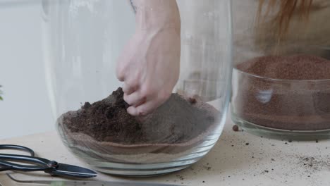 Female-botanist-creates-a-small-ecosystem-in-a-glass-terrarium-and-puts-the-soil-layer-for-a-live-tiny-environment-concept-close-up