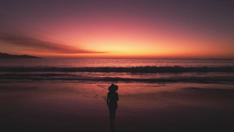 Aerial-Drone-4K-Flyover-Woman-Silhouette-Walking-Towards-Ocean-With-Bright-Red-Sunset