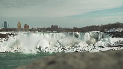 Wide-view-of-icy-Niagara-Falls-in-winter,-reveal-pan-from-behind-wall