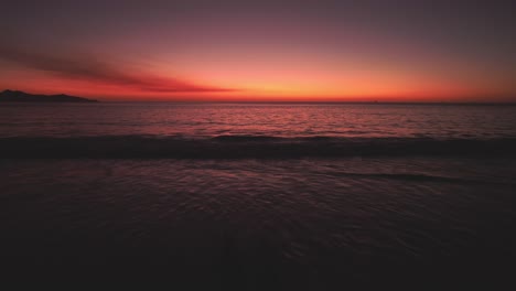 Romantische-Paarsilhouette-Mit-Blick-Auf-Strandwellen-Und-Leuchtend-Roten-Sonnenuntergang-Am-Horizont,-4K-Drohne