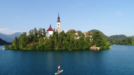Isla-Bled-Con-La-Iglesia-Y-La-Gente-Remando-En-El-Lago-Azul,-Pequeño-Paraíso-Natural-De-Eslovenia,-Vista-Aérea