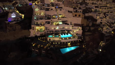 Aerial-View-of-Luxury-Santorini-Island-Resort-at-Night,-Lights-on-Swimming-Pool-and-White-Buildings