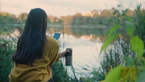 Young-woman,-long-hair,-near-lake-at-sunrise,-on-phone-with-thermos-in-hand