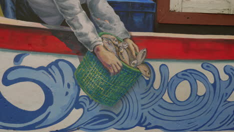Fisherman-catching-fish-in-a-bucket,-close-up-detail-of-wall-art-in-Asia
