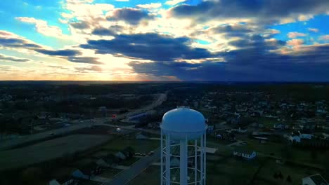Aerial-orbit-of-water-tower-in-suburban-neighborhood-revealing-a-stunning-sunset