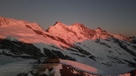 Luftaufnahme-Eines-Gebäudes-Auf-Den-Alpen,-Sonnenaufgang,-Orange-Beleuchtete-Felsige-Berge