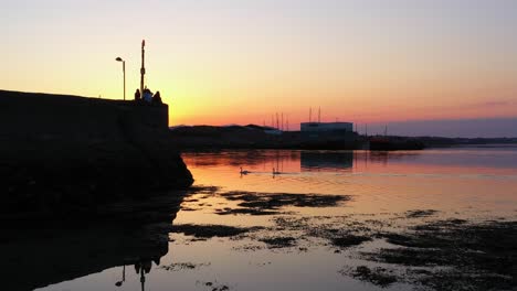 Schwäne-Gleiten-über-Ruhiges-Wasser,-Atemberaubende-Rosa-Und-Orangefarbene-Sonnenaufgangsreflexionen,-Einheimische-Genießen-Die-Aussicht-Am-Nimmos-Pier-In-Galway,-Irland