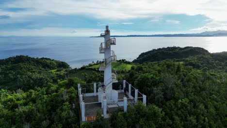 Rotating-Aerial-View-of-White-Lighthouse-atop-mountain-in-lush-rainforest-with-vast-ocean-and-cloud-sky-in-background
