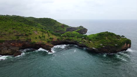 Cliff-covered-in-tropical-trees-extending-over-ocean-as-waves-crash-below,-Indonesia