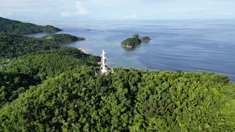 Stunning-Aerial-View-of-white-Bote-Lighthouse-atop-lush-tree-covered-Mountains-revealing-tropical-coastline-and-stunning-ocean-bay