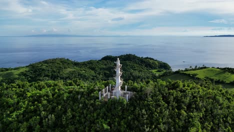 Aerial-view-zooms-out-from-Bote-Lighthouse-atop-a-tree-covered-hill-in-Catanduanes,-Philippines,-revealing-the-entire-island-and-ocean