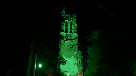 Beaumont-Tower-on-the-campus-of-Michigan-State-University-lit-up-at-night-in-green-in-honor-of-the-victims-of-the-February,-2023-mass-shooting-with-gimbal-video-walking-by-trees-forward