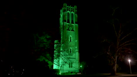 Beaumont-Tower-on-the-campus-of-Michigan-State-University-lit-up-at-night-in-green-in-honor-of-the-victims-of-the-February,-2023-mass-shooting-with-ws-walking-forward-through-trees