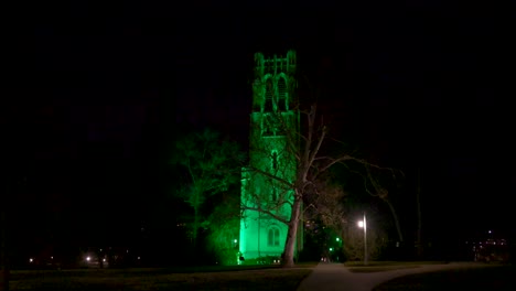 Beaumont-Tower-on-the-campus-of-Michigan-State-University-lit-up-at-night-in-green-in-honor-of-the-victims-of-the-February,-2023-mass-shooting-with-wide-video-walking-on-sidewalk