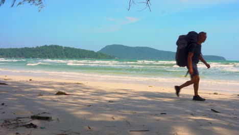 male-and-female-tourist-walk-along-tropical-beach-carrying-rucksacks,-islands-and-sea-in-background