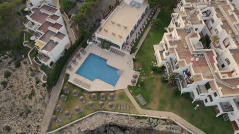Aerial-view-descending-Birdseye-over-Mallorca-holiday-resort-on-Mediterranean-coastal-cliff-edge-to-clear-blue-pool