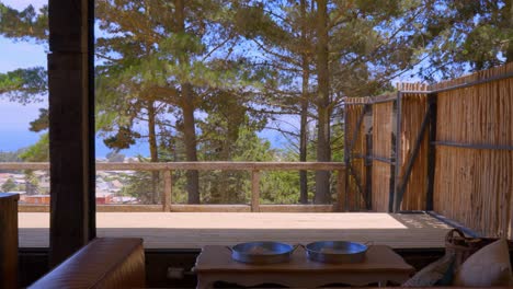 Recording-towards-the-outside-of-a-house,-you-can-see-a-sofa-and-in-the-background-a-rustic-terrace-with-a-pine-forest,-daylight-with-a-blue-sky