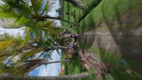 Vertical-FPV-drone-following-shot-of-blond-woman-driving-on-a-motorcycle-on-a-gravel-road-between-palm-trees