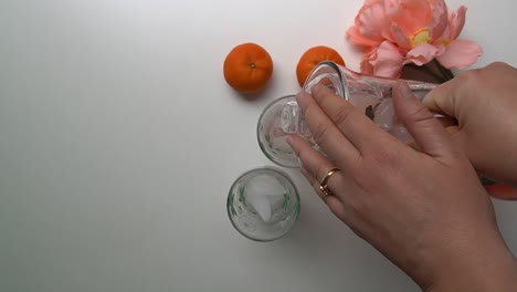 Woman-is-pouring-ice-tea-in-glasses,-top-fixed-view,-flower-plant-and-two-mandarin-oranges-located-near-the-glasses