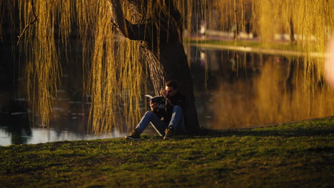 Young-man-reading-a-book-while-sitting-under-a-golden-tree-by-a-public-city-park-lake,-at-dusk