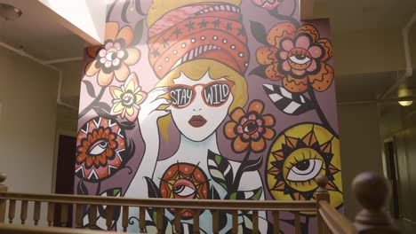 "Stay-Wild"-Colorful-Mural-with-Sun-Shining-indoors,-Woman-wearing-large-hat-around-flowers---Slow-Motion