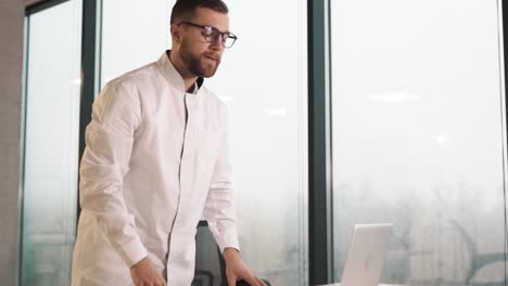 Focused-male-doctor-sits-at-a-desk-in-a-modern-clinic-with-panoramic-windows,-looks-at-a-laptop-and-reflects