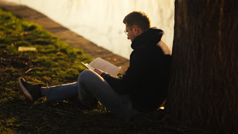 Slow-motion-shot-of-a-man-relaxing-reading-his-book-at-the-base-of-a-tree-beside-a-lake